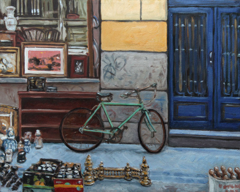 vintage green bike leaning against a wall with graffiti, between a dark blue door and a store window, and amidst other flea market items: guages in boxes, brass grate, bowling pins, glasses, vases, and a wooden cupboard